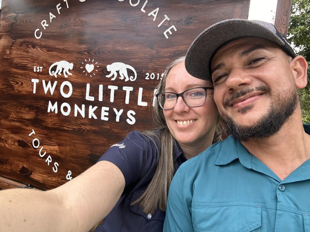 Lindy and Ismael smiling in a selfie in front of the Two Little Monkeys wooden sign